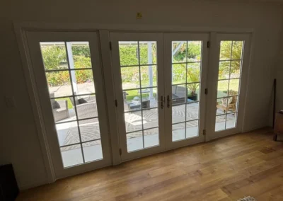 Interior view of French doors leading to a patio with outdoor furniture and lush greenery visible beyond. Warm wooden floor and rug.