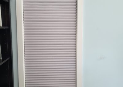 A window with white trim features vertical, light purple blinds against a pale blue wall, with a dark furniture piece below.