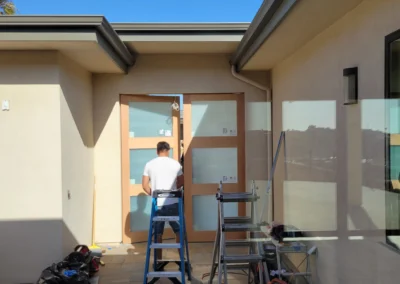A worker on a ladder installs a door frame, surrounded by tools and drop cloths, under a clear blue sky.