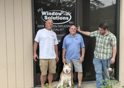 Three men in casual attire stand outside a storefront with a sign reading "My Window Solutions," accompanied by a friendly dog.